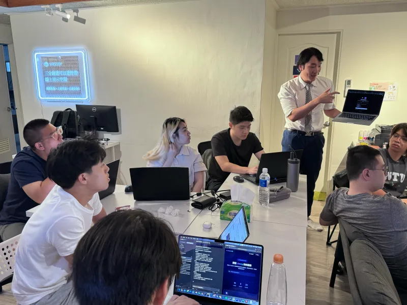 講師在辦公室對一桌學員講解 AI 工作流,桌上多台筆電同時操作