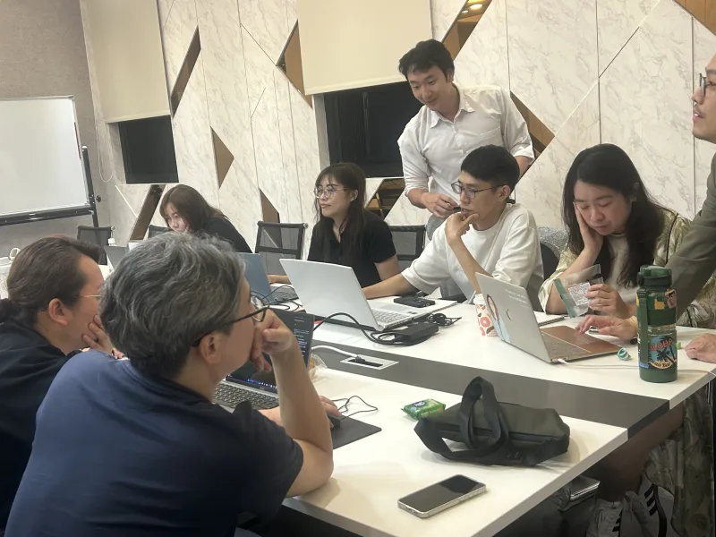 講師彎身在長桌旁為學員解答,會議室內多位學員同時實作