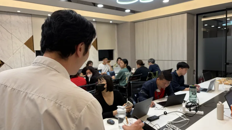 講師從教室後方觀察學員操作,長桌兩側坐滿參與者